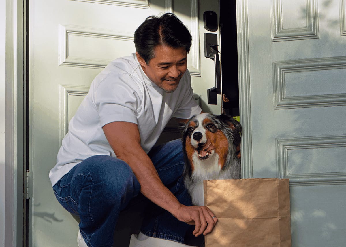 A man crouches at his front door to greet his Australian shepherd
